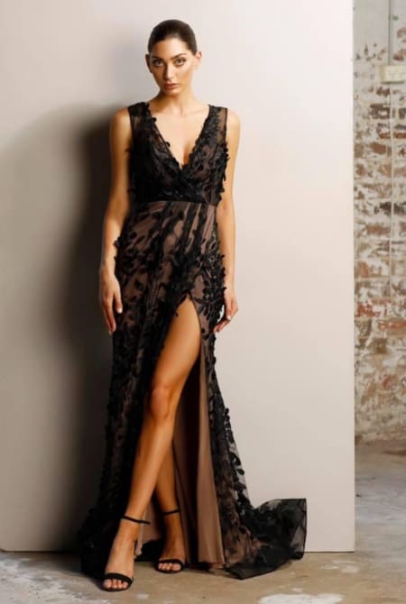 A woman poses confidently against a minimalist wall, dressed in the ORNELA JX1019 sheer black evening gown adorned with intricate floral designs. The gown boasts a high slit that reveals one leg. She finishes off her look with black high-heeled sandals and has neatly pulled back her dark hair. Angels Formal Wear