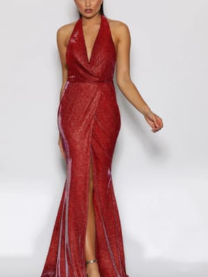 A woman stands wearing a shimmering red TIARNA JX2108 halter neck gown that features a plunging neckline and high slit. Her hair is slicked back, and she poses with one hand on her hip, against a plain white background. She wears high-heeled shoes. Angels Formal Wear