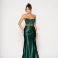 A woman wearing a strapless emerald green gown stands against a plain white background. The dress features a beaded bodice with sheer details and a satin skirt with a high slit. She has her hair styled in an updo and is smiling softly with one hand on her hip. Angels Formal Wear