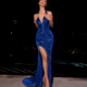 A woman standing confidently on a tiled surface against a night backdrop is wearing the AZALEA NC2022, a shimmering blue halter-neck gown with a high slit. With her hands near her shoulders, she showcases the dress's elegant design and pairs it with sparkling high heels, creating an image reminiscent of an AZALEA event. Angels Formal Wear