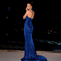 A woman stands on a tiled floor in a long, shimmering blue AZALEA NC2022 gown with a train. The gown features a halter neckline and an open back. She poses gracefully with her hand near her face, while the dark background with faint lights in the distance complements the NC2022 vibe perfectly. Angels Formal Wear