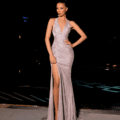 A woman stands confidently on a dimly lit pathway at night, wearing the shimmering, form-fitting AZALEA NC2022 gown with a high slit. Her hair is styled in an updo. The background is dark, with a faintly lit structure visible; the scene evokes the ambiance of an AZALEA festival night. Angels Formal Wear