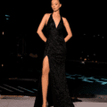 A woman stands confidently in the elegant AZALEA NC2022 evening gown, featuring a deep V-neckline and high slit that reveals her left leg. She wears high heels and poses outdoors with a pool and city lights in the background. The sophisticated nighttime setting perfectly complements her look at the NC2022 event. Angels Formal Wear