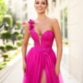 A woman in a vibrant pink, strapless gown stands outdoors with a smile. The AURORA TK554 dress she wears is adorned with intricate floral embellishments and features a high front slit, exuding elegance. In the background, there is a well-lit street lined with greenery and a light-colored building. Angels Formal Wear