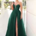 A woman stands outdoors wearing an elegant, strapless, dark green gown with a deep V-neckline and a high slit. She holds a small blue flower and smiles, her AURORA TK554-like glow contrasting beautifully against the blurred buildings and greenery in the background. Angels Formal Wear