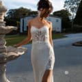 A woman in the ASTRID PO970 gown, featuring a white sequin fabric with a floral design and a thigh-high slit, poses outdoors near a decorative fountain. She is looking down, her updo hairstyle adorned with white flowers, embodying the elegance of the dress. In the background, a large house and trees are visible. Angels Formal Wear