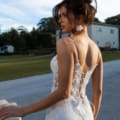 A woman wearing the ASTRID PO970 dress, characterized by its intricate design, thin straps, and lace-up back, stands outdoors. Elegance radiates from her pose as she gazes to the side. Her hair is styled in an updo with loose strands. The background showcases green grass, trees, and buildings. Angels Formal Wear