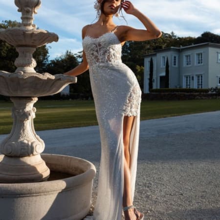 A woman stands next to an ornate stone fountain outdoors, wearing a shimmering, sleeveless, high-slit gown from the ASTRID PO970 collection. She has one hand on her forehead and the other resting on the fountain. In the background is a large, light-colored house surrounded by greenery. Angels Formal Wear