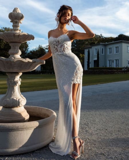 A woman stands next to an ornate stone fountain outdoors, wearing a shimmering, sleeveless, high-slit gown from the ASTRID PO970 collection. She has one hand on her forehead and the other resting on the fountain. In the background is a large, light-colored house surrounded by greenery. Angels Formal Wear