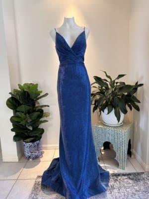 A shimmering blue floor-length gown known as the NEW YORK TH029 is displayed on a mannequin. Featuring a deep V-neck with thin straps, a fitted bodice that wraps at the waist, and a flowing skirt, this dress exudes elegance. Behind the gown are potted plants and a small table with a white pot on it, evoking chic New York style. Angels Formal Wear