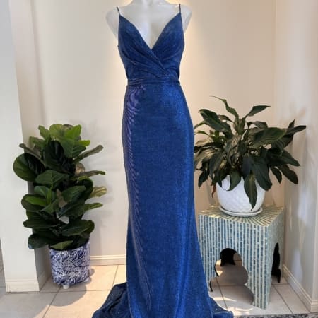 A shimmering blue floor-length gown known as the NEW YORK TH029 is displayed on a mannequin. Featuring a deep V-neck with thin straps, a fitted bodice that wraps at the waist, and a flowing skirt, this dress exudes elegance. Behind the gown are potted plants and a small table with a white pot on it, evoking chic New York style. Angels Formal Wear