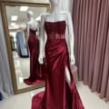 A white mannequin showcases the ONDINE PO2444 strapless evening gown in burgundy, featuring a beaded bodice and a high side slit. The floor-length gown includes a slight train. Positioned behind the mannequin, a large mirror reflects the dress from both back and side angles, accentuating its sophisticated design. Angels Formal Wear