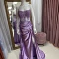 A mannequin is dressed in the ONDINE PO2444 lilac satin evening gown with a lacy, strapless bodice adorned with floral appliqués. The floor-length skirt features a high slit, and the ensemble is displayed in a fitting room with mauve curtains and a gold-framed mirror. Angels Formal Wear