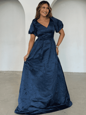 A woman stands indoors, smiling while posing in a dark navy blue, floor-length SOPHIA PUFF SLEEVE MAXI - TL evening gown with short puff sleeves and a V-neckline. The fabric has a subtle marbled pattern. She holds a portion of the dress in one hand, and the background is a plain, light-colored wall with two arched alcoves. Angels Formal Wear