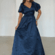 A woman stands indoors, smiling while posing in a dark navy blue, floor-length SOPHIA PUFF SLEEVE MAXI - TL evening gown with short puff sleeves and a V-neckline. The fabric has a subtle marbled pattern. She holds a portion of the dress in one hand, and the background is a plain, light-colored wall with two arched alcoves. Angels Formal Wear