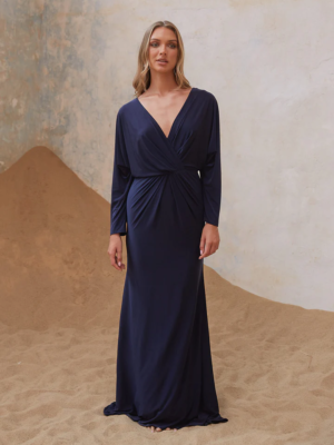 A woman stands on a sandy surface, wearing the ADAIR TO2436 long, navy blue dress with a deep V-neckline and gathered twist detail at the waist. The background features a faded, textured wall with hints of blue and beige tones. The dress has long sleeves and flows to the ground. Angels Formal Wear