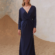 A woman stands on a sandy surface, wearing the ADAIR TO2436 long, navy blue dress with a deep V-neckline and gathered twist detail at the waist. The background features a faded, textured wall with hints of blue and beige tones. The dress has long sleeves and flows to the ground. Angels Formal Wear