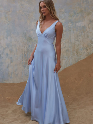 A woman stands on a sandy surface wearing the light blue, floor-length, sleeveless AVONLEA TO2428 dress. The background features a softly textured, pastel-colored wall. She gazes forward with a relaxed expression, holding the skirt of her dress lightly with one hand. Angels Formal Wear