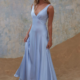 A woman stands on a sandy surface wearing the light blue, floor-length, sleeveless AVONLEA TO2428 dress. The background features a softly textured, pastel-colored wall. She gazes forward with a relaxed expression, holding the skirt of her dress lightly with one hand. Angels Formal Wear