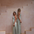 Two women with long, wavy hair stand back to back on a staircase against a soft peach-colored backdrop, both elegantly dressed in sleeveless, floor-length pastel green dresses with side slits from the CARY TO2432 collection. Their sophisticated attire enhances the scene's timeless charm. Angels Formal Wear