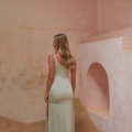 A woman with long, wavy blonde hair is standing with her back to the camera, wearing a light green sleeveless dress with a side slit. She is in a room with textured, earth-toned walls and a sand-covered floor, creating an ambiance reminiscent of the CARY TO2432 deserts. Angels Formal Wear