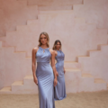 Two women stand in front of a beige, stepped wall. They both wear floor-length, silky lavender dresses from the CORDELIA TO2433 collection. The woman in front has her hands at her sides and faces the camera, while the woman behind is slightly turned with her hands behind her back. The floor appears to be sand. Angels Formal Wear