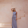 A woman in a long, light blue satin dress, identified as the CORDELIA TO2433, stands on a pastel-colored staircase. She poses with one hand resting on her head against a backdrop of soft, warm tones and minimalistic design. Palm leaves slightly enter the frame from the top right corner. Angels Formal Wear