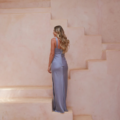 A woman with long, flowing blonde hair stands on a set of light peach-colored, geometric stairs, her back turned to the camera. She is wearing the CORDELIA TO2433, a form-fitting lavender gown featuring a thigh-high slit and an elegant floor-length design. The minimalist setting perfectly complements the serene vibe of the dress. Angels Formal Wear