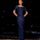 A woman stands confidently at night in front of a dimly lit background. Wearing the fitted, floor-length, short-sleeved blue LUXETTE NC2002 dress adorned with a grid pattern and sequins, her hair is styled in a sleek updo, and she has a serious expression. Angels Formal Wear