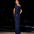 A woman is standing near a pool at night, facing away from the camera and looking to her left. She is wearing a form-fitting, navy blue LUXETTE NC2002 dress with geometric patterns. The background is dark, with city lights faintly visible. Angels Formal Wear