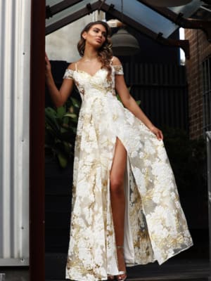 A woman stands confidently in a flowing white dress with gold floral patterns, the SHANIKA JX3062 - SALE. The dress features off-the-shoulder sleeves and a high slit on one side, revealing her leg. She is under a transparent, angled roof structure, next to a metal column and wooden plants. Angels Formal Wear