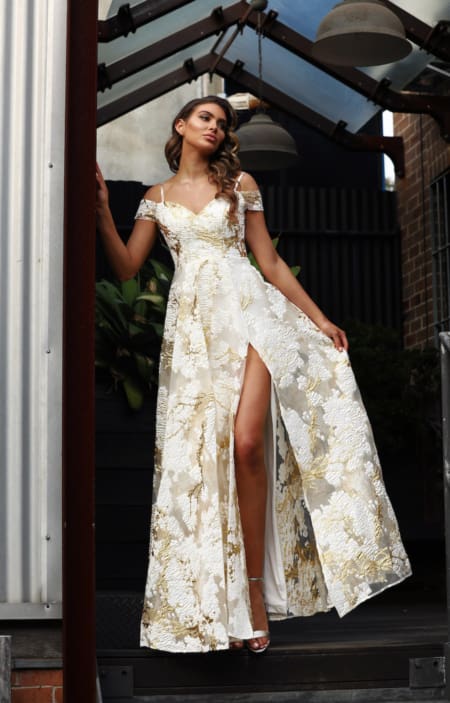 A woman stands confidently in a flowing white dress with gold floral patterns, the SHANIKA JX3062 - SALE. The dress features off-the-shoulder sleeves and a high slit on one side, revealing her leg. She is under a transparent, angled roof structure, next to a metal column and wooden plants. Angels Formal Wear