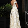 A woman wearing the elegant SHANIKA JX3062 off-shoulder white gown with gold floral patterns stands outdoors, facing away. She is looking back over her shoulder, and the dress, currently on sale, features a low-cut back. She is positioned beside large potted plants and industrial-style railings. Angels Formal Wear