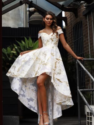 A woman stands on stairs, elegantly dressed in an off-shoulder, high-low SHONTINA JX dress adorned with a white and gold floral pattern. She gently lifts the side of her dress to reveal her silver high-heeled sandals. Her long, wavy hair cascades down her back as she gazes serenely into the distance, with lush green plants providing a picturesque backdrop. Angels Formal Wear