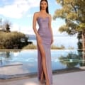 A woman stands by a pool with a scenic backdrop of trees and a lake. She is wearing the ISABELLA NC2010, a strapless, form-fitting lavender dress with a high slit, paired with silver high-heeled sandals. The sky is partly cloudy, featuring blue tones that complement the serene environment. Angels Formal Wear