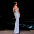 A woman stands elegantly by a dimly lit pool at night, wearing the long, shimmering STEVIE NC2037 gown. Her back is slightly turned to the camera, showcasing the gown's intricate detailing and her graceful pose. The scene exudes sophistication and glamour, perfectly encapsulating the essence of STEVIE chic. Angels Formal Wear