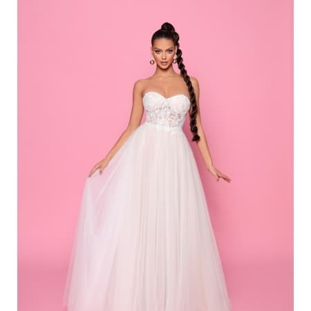 Against a pink background, a woman showcases the SUZIE NP147W ensemble, a strapless white gown featuring a sheer lace bodice and flowing tulle skirt. She wears her long braided hair styled to one side and accessorizes with large hoop earrings. With arms softly extended holding the dress, she exudes effortless elegance. Angels Formal Wear