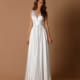 A woman stands against a plain brown background wearing the SKYLA NBM1013, a floor-length, sleeveless white gown featuring a deep V-neck and a cinched waist. Her long, wavy hair cascades down her shoulders as she maintains a neutral expression. Angels Formal Wear