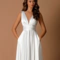 A woman with long wavy hair wears a SKYLA NBM1013 sleeveless, floor-length white gown featuring a deep V-neck and a cinched waist. She stands against a plain brown background with her hands in the dress pockets, looking slightly to the side. Angels Formal Wear