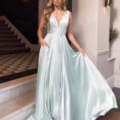 A woman in a light blue, satin, floor-length gown labeled SKYLA NBM1013 stands on steps indoors. The dress features a plunging neckline and pockets. With long, wavy blonde hair, she holds part of her gown in one hand, letting it flow elegantly around her. Angels Formal Wear