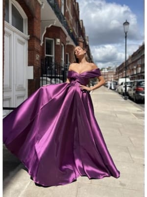 A woman in an elegant WILLOW JX6027, an off-shoulder, flowing purple gown, stands on a sidewalk in front of a row of red brick buildings and parked cars. She poses confidently with her gown spread out like the branches of a willow tree and gazes upward, capturing a sense of glamour and grace. Angels Formal Wear