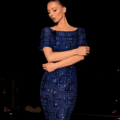 A woman wearing the form-fitting LUXETTE NC2002, an off-the-shoulder blue sequin dress adorned with a geometric pattern, stands with her arms crossed. She poses against a dark, outdoor backdrop with her hair styled in an updo. Her LUXETTE ensemble embodies elegance and sophistication amidst the night. Angels Formal Wear