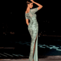 A person, captured from behind, exudes the elegance of the LUXETTE NC2002 in a form-fitting light green gown adorned with a geometric pattern and featuring a high slit. They stand on a tiled surface against a dark, blurry backdrop that suggests nighttime. The individual touches their head with one hand, perfectly channeling the essence of LUXETTE NC2002. Angels Formal Wear