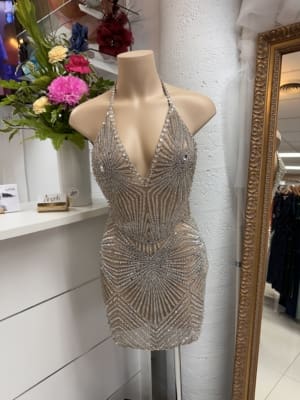 An AMARNI PORTIA AND SCARLET - SALE mannequin showcases a silver halter neck mini dress elaborately decorated with sequin patterns. The dress highlights a plunging neckline. In the background, a mirror, flowers, and other dresses are present in the store setting. Angels Formal Wear