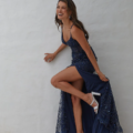 A woman stands in front of a white wall, smiling and posing with one leg bent at the knee. She is wearing a navy blue sequined gown with a high slit, revealing a pair of KAIRI PO2460 white high-heeled sandals. Her hair is styled loosely, and she appears joyful and confident. Angels Formal Wear