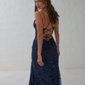 A woman with flowing brunette hair stands slightly turned to the side, adorned in a backless, navy blue sequined gown featuring intricate crisscross straps. She gazes over her shoulder against a simple, light-colored backdrop, epitomizing the elegance of the KAIRI PO2460. Angels Formal Wear