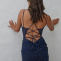 A woman with wavy brown hair stands with her back to the camera, facing a white textured wall with her hands resting on it. She is wearing a navy blue KAIRI PO2460 dress featuring thin straps and intricate lace detailing, complete with crisscross lacing at the back. Angels Formal Wear