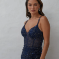 A woman stands against a plain, light-colored background wearing a dark blue KAIRI PO2460 sleeveless dress, adorned with sequins and beadwork. With long, wavy hair and drop earrings, she looks directly at the camera with a neutral expression. The KAIRI PO2460 design perfectly complements her elegant look. Angels Formal Wear