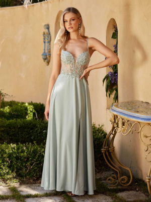 A woman stands outdoors against a decorative wall, wearing the MOSS PO24145, a long, light green dress with a fitted, semi-transparent bodice embellished with lace and beading. Her long blonde hair cascades gracefully as she poses with one hand on her hip. The surroundings feature lush greenery and hints of moss. Angels Formal Wear