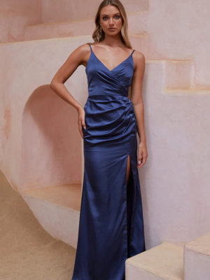 A woman stands on light-colored steps in a sophisticated setting, wearing a deep blue NEPTUNE TO2426 satin, floor-length gown with spaghetti straps, a ruched side, and a thigh-high slit. With one hand on her hip and an elegant, poised expression on her face, she perfectly showcases the style. Angels Formal Wear